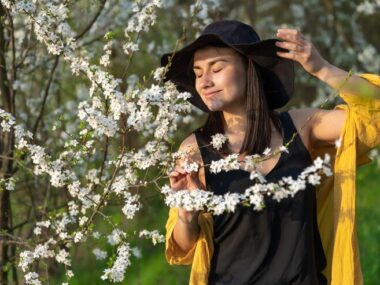 An attractive girl in a hat among blooming trees enjoys the smell of spring flowers