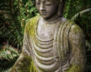 Vertical shot of a Buddha statue with moss on top and greenery on the distance