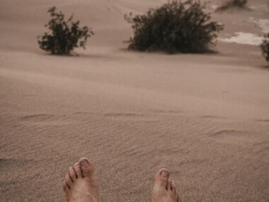 Male feet on soft desert sand sitting on a dune in desert at sunset with mountains and dramatic