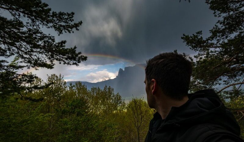 Man looking at rainbow by mountain against cloudy sky