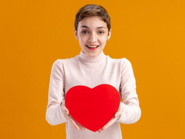 Young woman with short hair holding heart made of cardboard looking at camera happy and positivesmiling cheerfully valentines day concept standing over orange wall