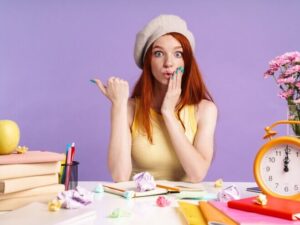 Photo of surprised student girl pointing finger at copyspace while sitting at desk with exercise books isolated over purple wall