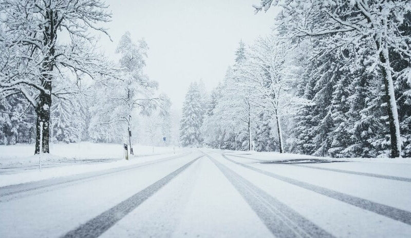 Wide shot of a road fully covered by snow with pine trees on both sides and car traces