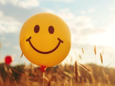 A yellow smiley face balloon floats in a field of tall grass with the sun shining brightly in the background